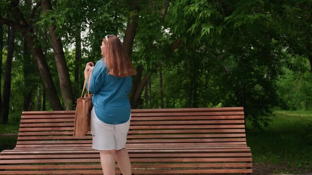 Rear view of woman with long brown hair in blue shirt and white shorts sitting on wooden bench in sunny park, removing items from bag, surrounded by fresh greenery