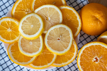 Sliced oranges and lemons on white plate with whole citrus fruits on checkered napkin close up top view. Concept of healthy eating, vitamin C nutrition, fresh fruits and natural immune boosting food.