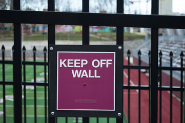 Maroon and white Keep Off Wall warning sign attached to a black metal fence at a sports stadium track