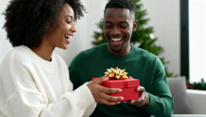 Happy African American couple with Christmas gift at home. Woman giving red present to surprised man in green sweater. Holiday celebration concept