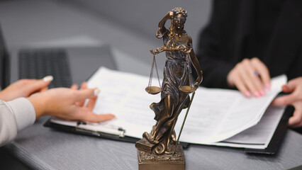 Bronze statue of Lady Justice holding scales, positioned on a desk with documents and a laptop,...