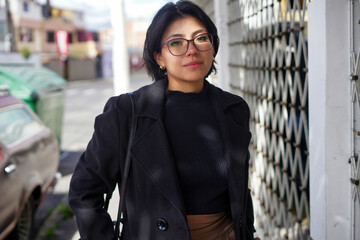 Young woman with short dark hair and glasses standing confidently on a city sidewalk, wearing a black coat and a black sweater