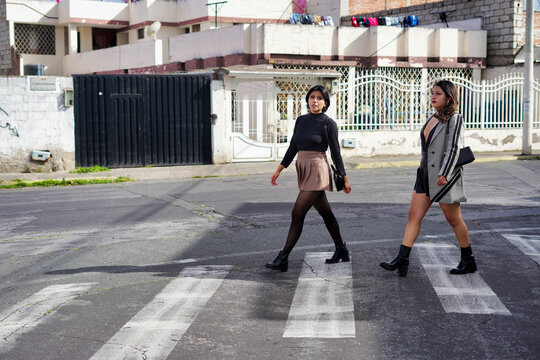 Two fashionable young women walking across a city crosswalk, side view full-length shot showing casual street-style outfits, - Powered by Adobe