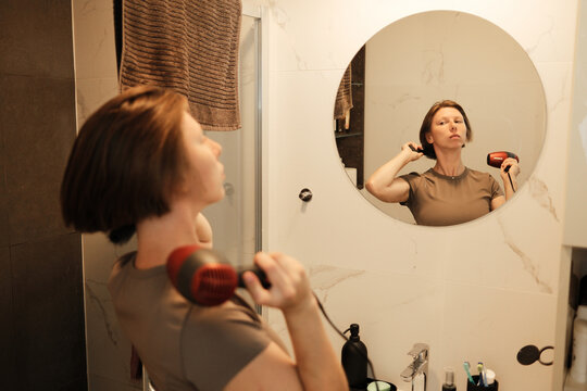 A woman dries her short hair with a hairdryer while looking into a bathroom mirror, focusing on her appearance and morning self-care ritual.