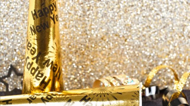 New Years Eve party decor on a twinkling gold and black background. Slow downward panning motion. Noisemakers, ribbon, confetti.