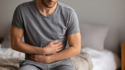 Unwell young man experiencing severe abdominal pain or stomachache, sitting on his bed feeling sick and uncomfortable
