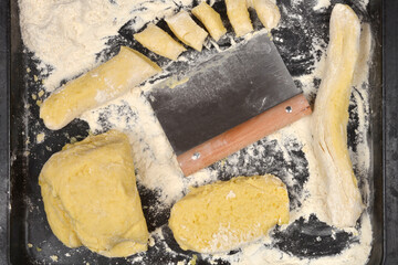 A bird's eye view showing different stages of gnocchi dough preparation, including a block, a rolled log, cut pieces, and a dough scraper tool on a floury dark tray