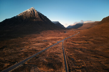 Naklejka premium Aerial view of Buachaille Etive Mor and the road through Glen Coe, Scotland. Dramatic mountain landscape, warm evening light, and vast moorland scenery in autumn. Natural landscape, no people