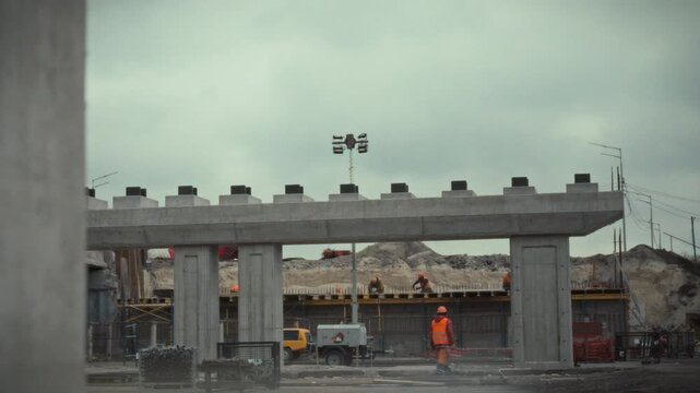 Construction workers are busy installing concrete bridge beams at a site. The work takes place under a cloudy sky, highlighting a collaborative effort in infrastructure development