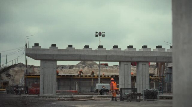 Workers are actively installing a concrete bridge at a busy construction site. Heavy machinery and building materials surround them as construction progresses in an urban environment during the day