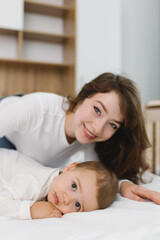 Cozy moment of a young woman and baby relaxing together at home, lying on a soft bed and smiling in natural light.