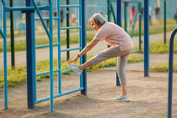Full body side view of an 87-year-old woman performing a hamstring stretch, touching her foot to a bar on a blue climbing ladder at an outdoor fitness area. Flexibility for seniors.