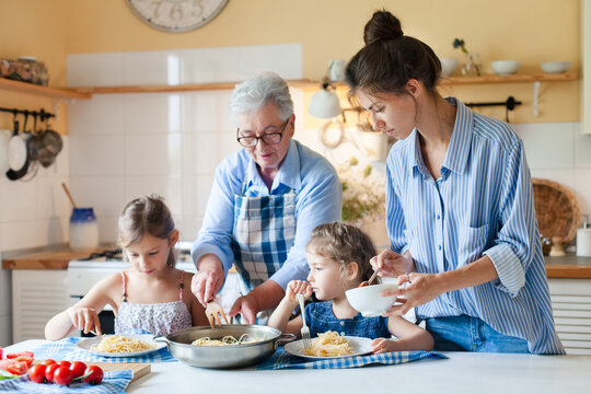 Multigenerational family cooking dinner and eating together at home kitchen, grandmother helping kids and mother feeding child with comfort food, pasta. Candid, authentic lifestyle moment - Powered by Adobe