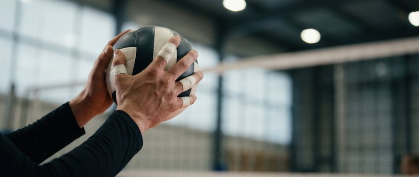 Male athlete preparing to serve a volleyball, gripping the ball tightly with hands wrapped in tape, showcasing focus and determination in a sports facility environment