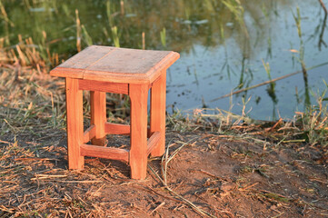 Small wooden stool stands on dry ground near calm water edge under warm sunlight. rustic handmade furniture placed in quiet natural environment. peaceful countryside atmosphere.