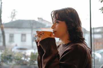pensive woman sipping coffee by window, holding warm mug and gazing at street view, soft morning light