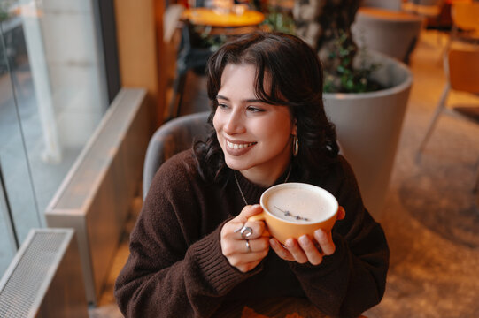smiling woman holding coffee cup indoors, warm cafe lighting, knit sweater, soft latte froth visible, potted