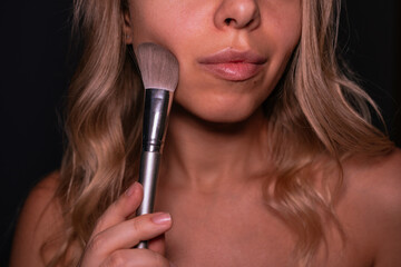 Close-up of a young blonde woman applying makeup with a soft brush, showing natural skin texture, glossy lips and a gentle beauty routine in a studio setting on a dark background