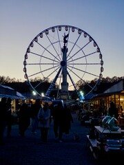 Twilight view of an illuminated Ferris wheel towering over a bustling outdoor vintage market with silhouetted people and antique displays in a charming evening ambiance