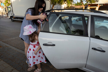 Mother preparing to put a bottle or sunscreen through the open back window of a white car while her daughter stands by the door.