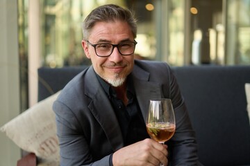 Older man sitting on coach drinking champagne based cocktail from wine glass, smiling.