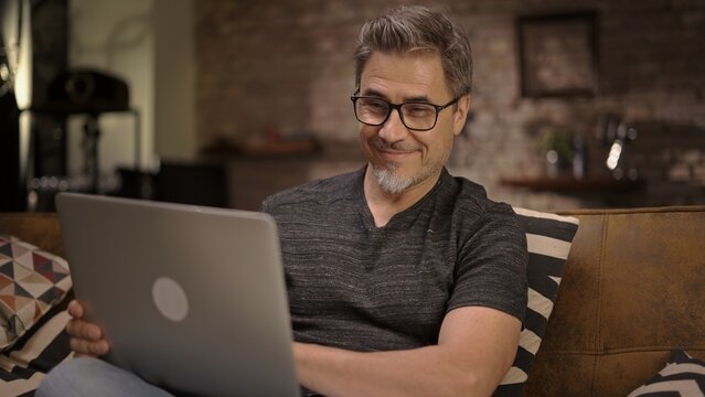 Older man working online with laptop computer at home sitting on couch in living room. Home office, browsing internet. Portrait of happy, mature age, middle age, mid adult man in 50s, smiling.