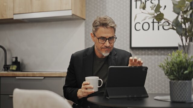 Businessman working with tablet computer in home office. Serious middle aged, mid adult, mature age man drinking coffee. Entrepreneur sitting at desk, managing business online.