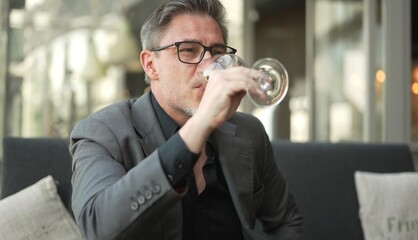 Older man sitting on coach drinking champagne based cocktail from wine glass, smiling.