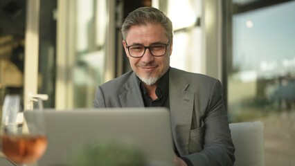 Happy businessman with laptop computer in restaurant. Middle aged man sitting at table in hotel lobby or business lounge in office, working, smiling.