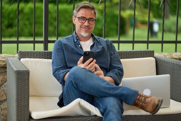 Man using mobile phone outdoor, sitting in garden at home, managing business online from home office. Focusing on work, thinking .