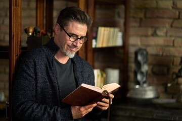 Happy older casual man standing by book shelf at home, reading, smiling. Portrait of mid adult, mature age man in glasses,