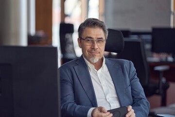 Business portrait - confident businessman sitting at desk working in office, using phone. Happy mid adult man in shirt and jacket, smiling. Bearded, glasses.