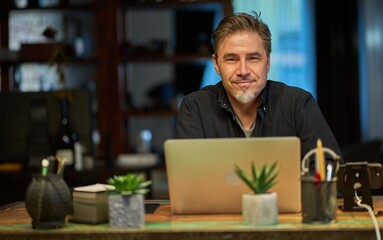 Older man sitting at desk in dark cosy room working on laptop computer in home office. Mature age, middle age, mid adult casual man in 50s, confident happy smiling.