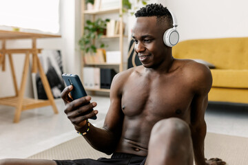 Young African man sitting on a mat, watching a workout video on his smartphone and wearing headphones at home