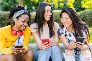 Three cheerful young women are enjoying a summer day outdoors, sitting together and sharing a laugh while using their smartphones