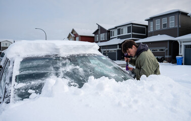Person scraping snow off car windshield after heavy snowfall in residential area.
