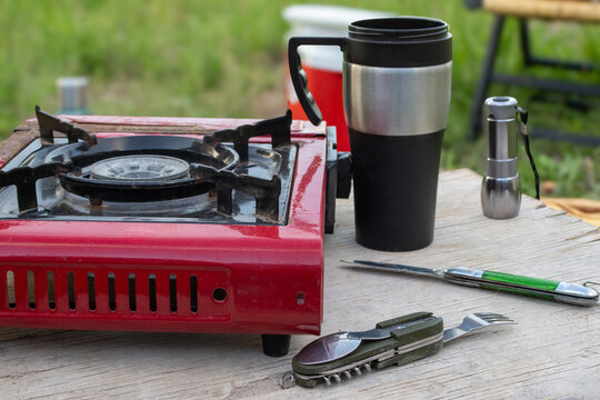 Portable gas stove, travel mug, flashlight and multi tool on a rustic wooden table, essential gear for outdoor cooking, camping picnics and relaxed days in nature