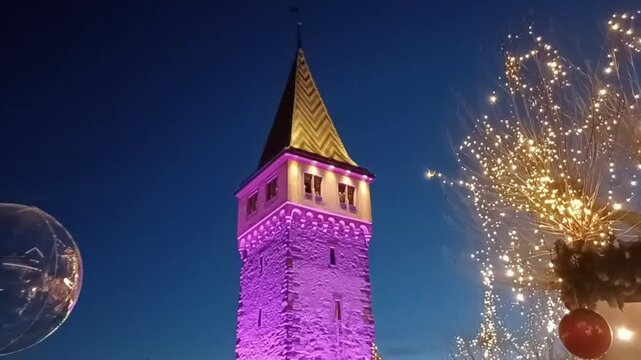Tilt up video shot of the illuminated Mangturm (Old Lighthouse) at the Christmas Market (Hafenweihnacht). The historic tower is lit in purple colors rising above festive wooden stalls at night