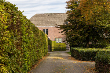 Brick house with arched window and tiled roof framed by tall hedges and autumn tree, gated driveway adds symmetry and privacy to this serene, well kept residential scene.