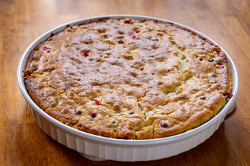 Freshly baked cranberry cake with a golden crust, cooling in a white rustic dish on a dark wood board. Homemade holiday dessert.