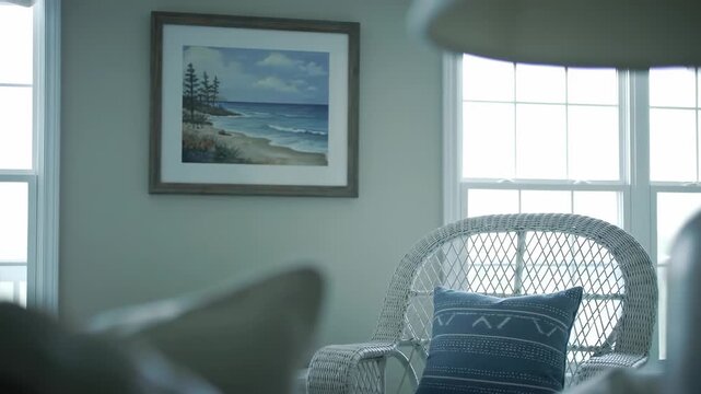 Beach painting hangs on wall next to window and white wicker chair with a blue patterned pillow in a softly lit room