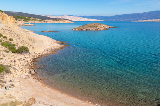 A secluded pebble beach meets the crystal clear turquoise water of the Adriatic Sea near Lopar. The sunny coastal landscape features rugged rocky terrain on the scenic Rab island in Croatia.