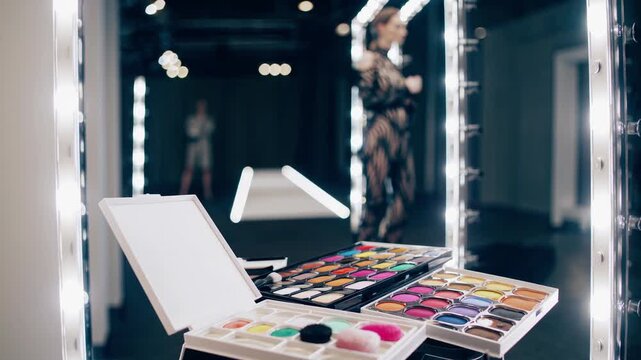 Professional makeup palette with various colorful eyeshadows and brushes placed on table in brightly lit dressing room with model reflected in mirror