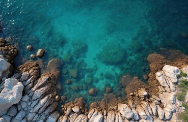 Aerial top view of clear blue sea water with submerged rocks and a rocky coastline. Waves gently lap against the shore creating a tranquil natural scene.