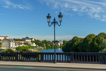 A street with a bridge and a river La Charente in city Saintes France. The street is lined with trees and houses. The sky is blue and clear