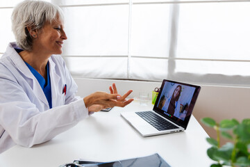 Mature adult doctor talking with female colleague through video call sitting on desk at medical consultation. E-learning, business and healthcare education concept