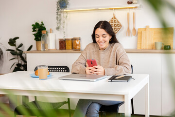 Young happy student woman using on mobile phone sitting on kitchen table at home, watching social media content or chatting online with friends
