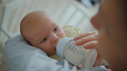 A mother is feeding a small baby formula from a bottle in front of a crib close up. The baby is eating with appetite and looking at her