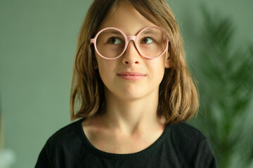 Fototapeta premium A small girl with round glasses looks sideways with sly squint and smile. Close up portrait showing emotions of curiosity, insight and childhood cleverness.
