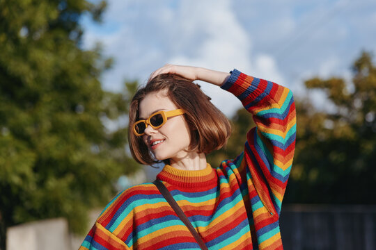 Young woman wearing a colorful striped sweater and yellow sunglasses, smiling with bob haircut in an outdoor portrait, casual fashion lifestyle pose in a sunlit park for travel editorial. - Powered by Adobe